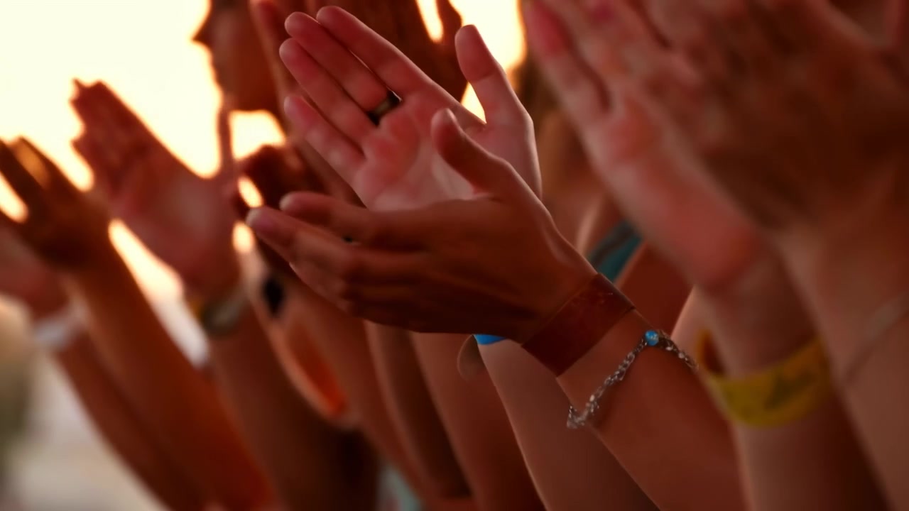 Multiple hands clap together in lively applause, captured from low side view with warm backlight, visible palm contact, finger motion, wristbands, jewelry, and soft blur conveying shared energy