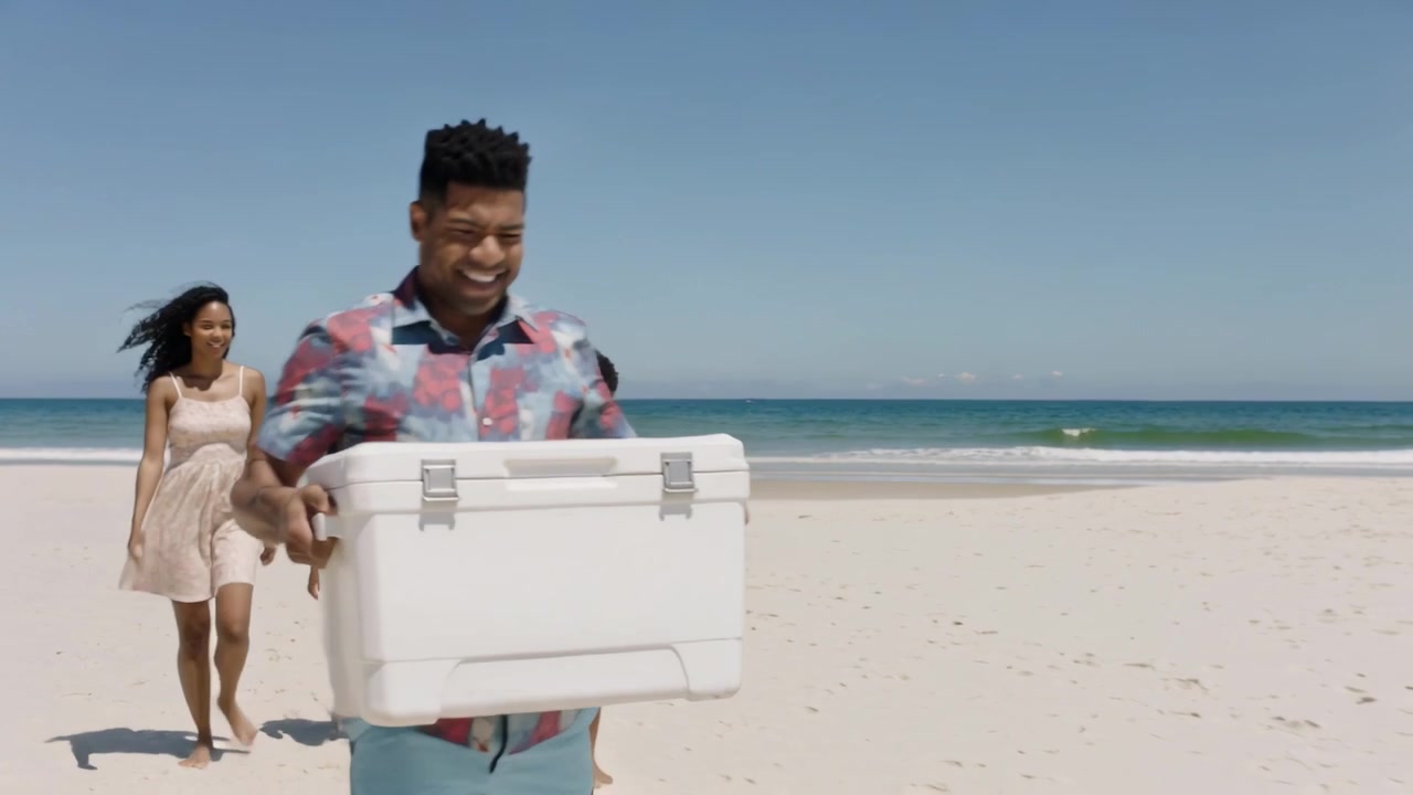 Walking man in patterned shirt carrying large white cooler on sandy beach, sunny sky drawing family