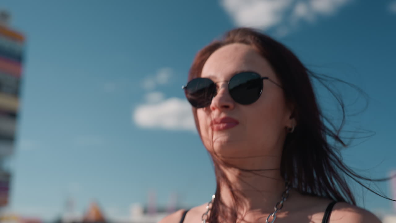 Side close up of woman in sunglasses and chain necklace walking outdoors under clear blue sky, showing confidence, urban lifestyle, individuality, and modern fashion attitude