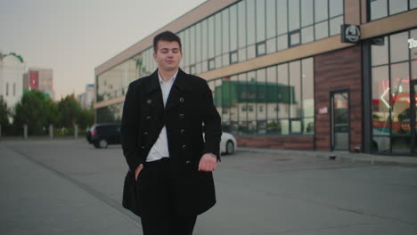 man in black coat adjusting coat, placing hand in pocket, walking majestically with office building and parked cars in background, confident, professional walk in urban business environment