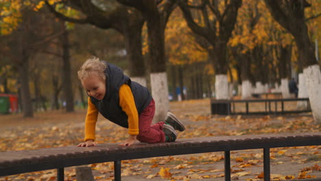 Un-Niño-Feliz-Se-Arrastra-En-Un-Banco-En-El-Parque-De-La-Ciudad-Caminando-El-Fin-De-Semana-De-Otoño.-Un-Niño-Sonriente-Disfruta-De-Una-Infancia-Despreocupada.-Un-Niño-Travieso-Y-Lindo.