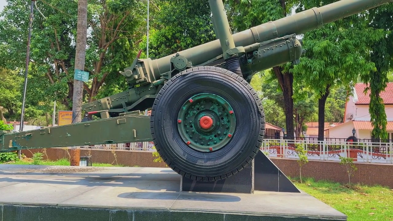 Ripresa panoramica cinematografica laterale di un cannone da campo indiano da 105 mm esposto su una piattaforma monumentale con grandi ruote, corpo militare verde e alberi in piena luce diurna.