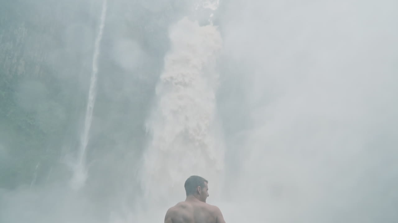 Man stands in front of crashing waterfall in rain forest surrounded by spray