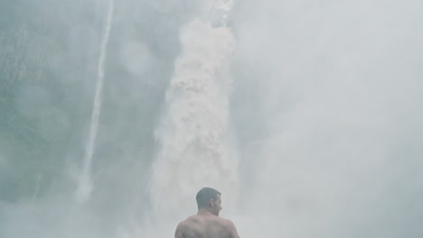 man stands in front of crashing waterfall in rain forest surrounded by spray