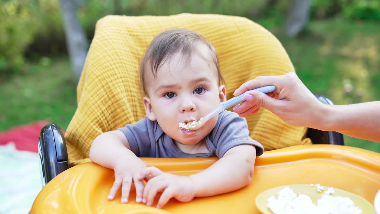 Caring mother giving full spoon to a cute adorable baby. Lovely little boy eating slowly looking at something attentively. Blurred nature backdrop.