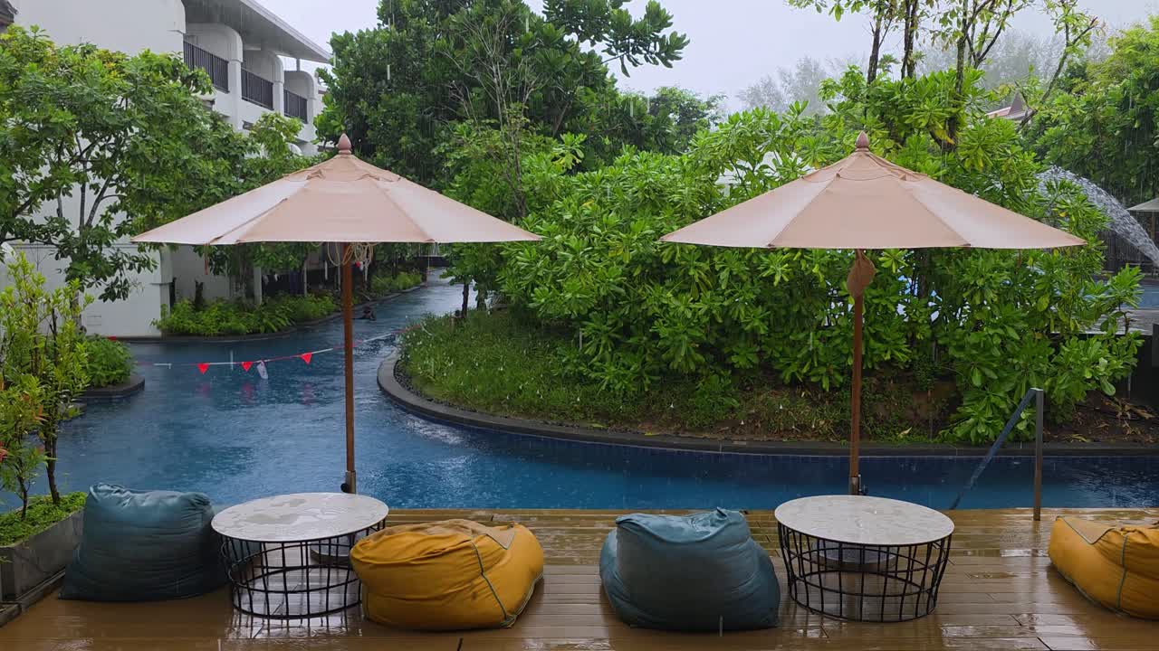 Wet wooden deck with colorful bean bag chairs and patio umbrellas overlooks curved pool surrounded by lush tropical garden during monsoon rain at JW Marriott Khao Lak Resort