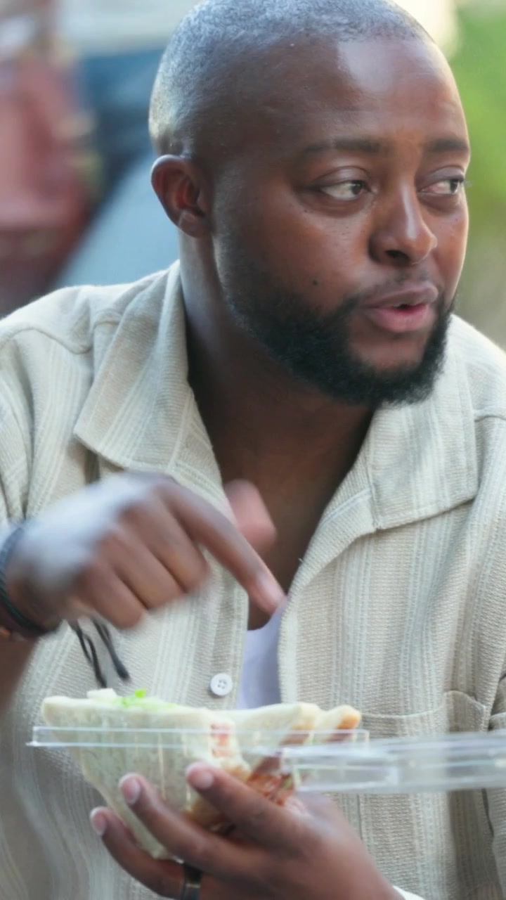 Vertical video: Panning camera showing man in shirt eating clear box on bench, reacting to passerby