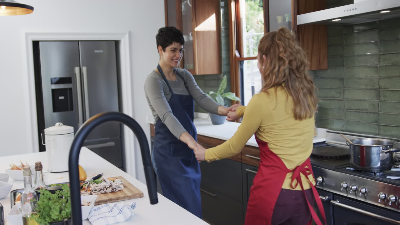 Feliz pareja de lesbianas caucásicas preparando comida, bailando y usando una tablet en una cocina soleada