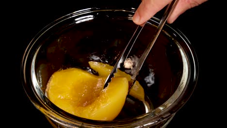 hand uses metal tongs to pick up a canned peach slice from a clear glass bowl on a black background, under bright studio lighting