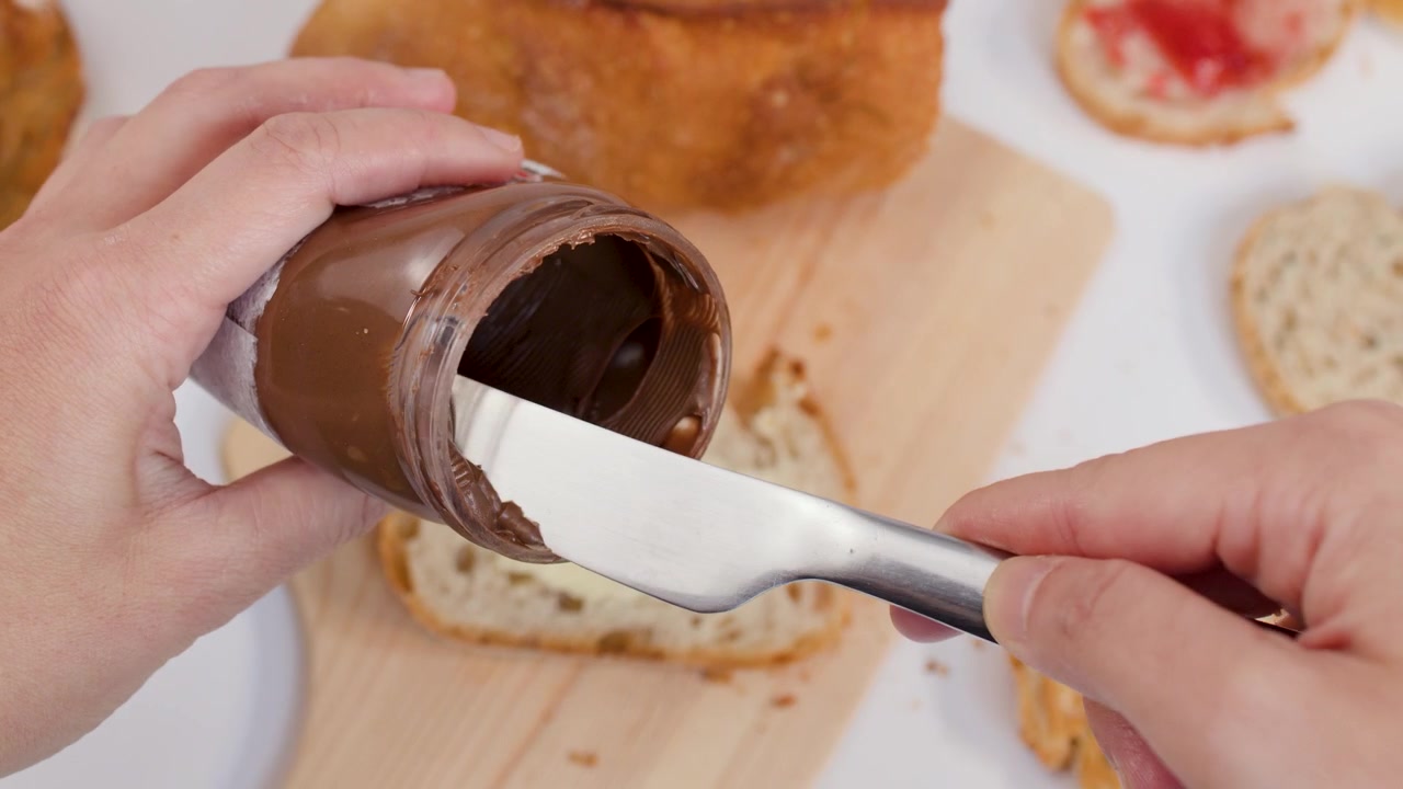 Mani usano un coltello di metallo per prelevare crema spalmabile al cioccolato da un vasetto di vetro.