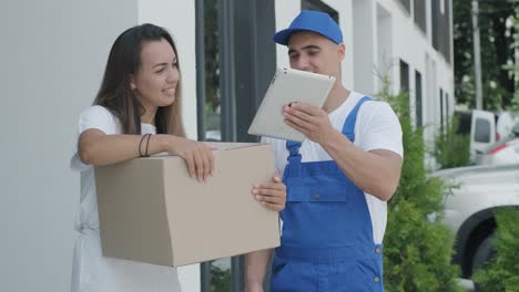 una mujer hermosa conoce a un repartidor que le da una caja de paquetes al lado de su casa, ella firma una firma electrónica en una tableta. concepto de mensajero, entrega a domicilio, compras en línea