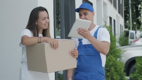 beautiful woman meets a delivery man who gives her a parcel box beside her home, she signs electronic signature on tablet. concept of courier, home delivery, online shopping