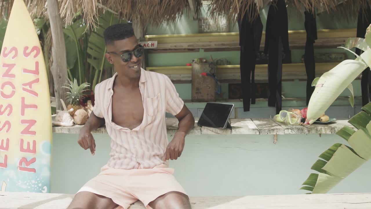 Glad afroamerikansk mand med solbriller slapper af uden for en surfbrætudlejningshytte, slow motion