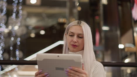 shoulder view of impulse buyer seated on bench by glass railing under sparkling bokeh lights, her focus on touchscreen tablet as she taps and smiles in cozy sweater capturing urban tech leisure moment