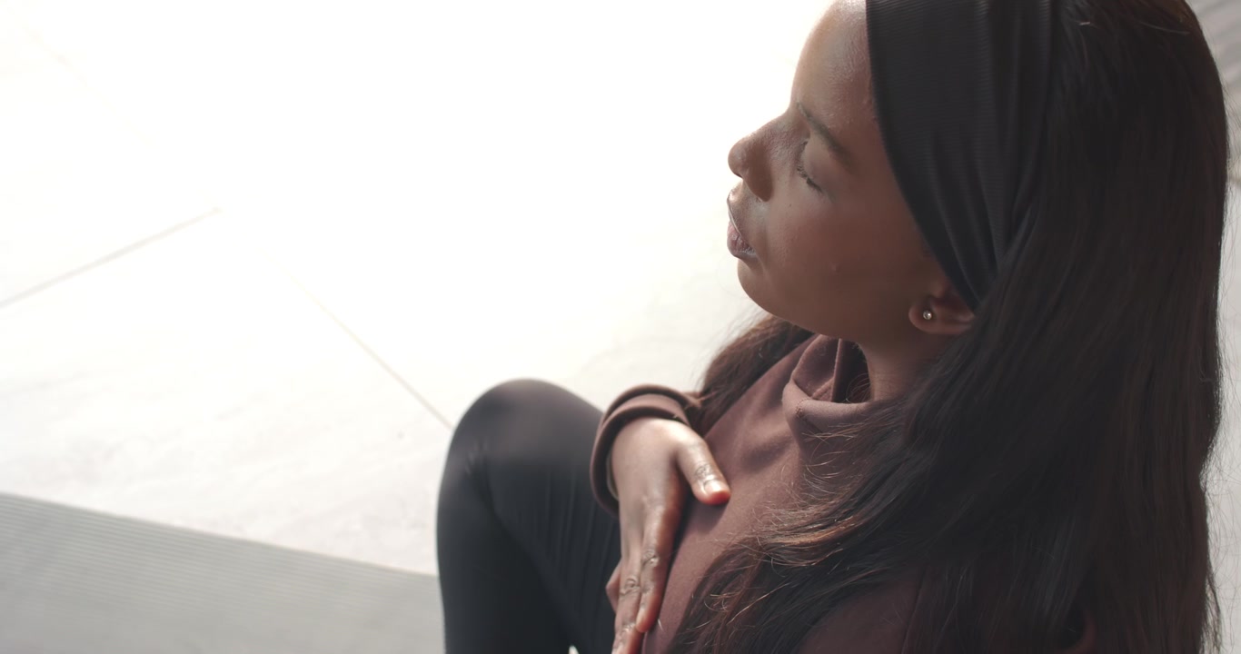 African American woman bringing hand to chest, practicing breathwork on mat in bright room
