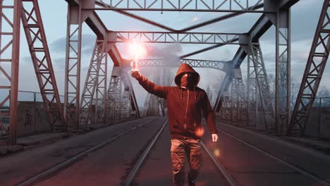 young man in hoodie and balaclava with red burning signal flare on the road under an old steel frame bridge, slow motion