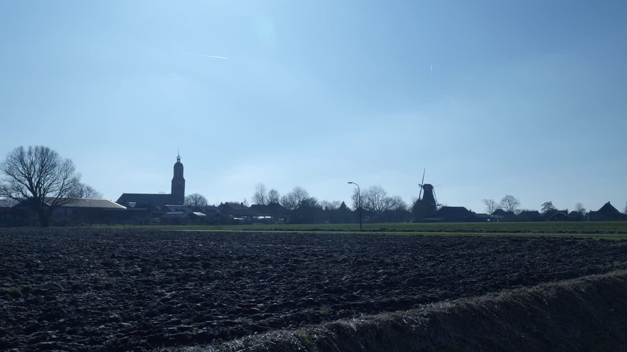 Landelijk dorpsgezicht met kerk en windmolen bij geploegd veld.