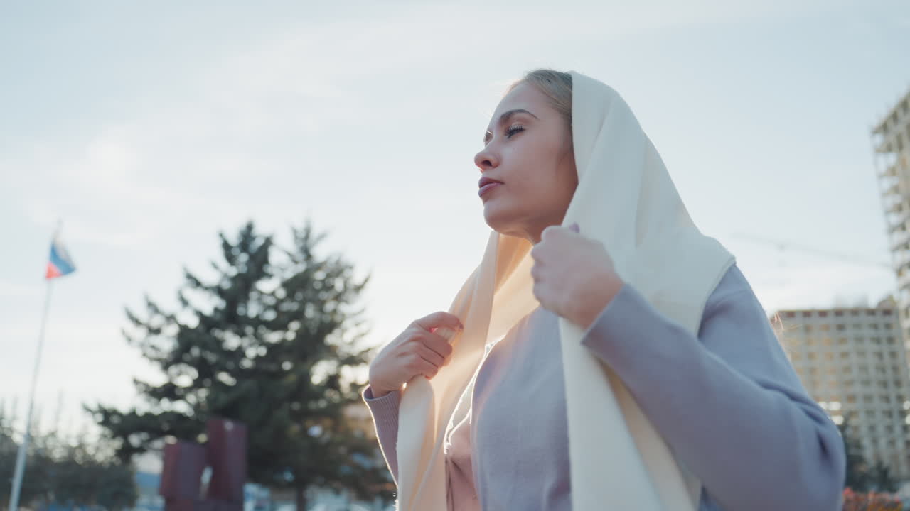 Gün batımında dışarıda krem rengi eşarp takan genç bir kadın, yumuşak arka ışığa karşı eşarbını düzeltiyor; arka planda şehir silüeti ve modern binalar, hafif esinti kumaşı hareket ettiriyor, düşünceli ifade.