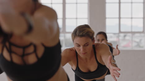 hermosa mujer caucásica de yoga practicando meditación en pose guerrera con un grupo de mujeres multirraciales disfrutando de un estilo de vida saludable haciendo ejercicio en un gimnasio al amanecer.