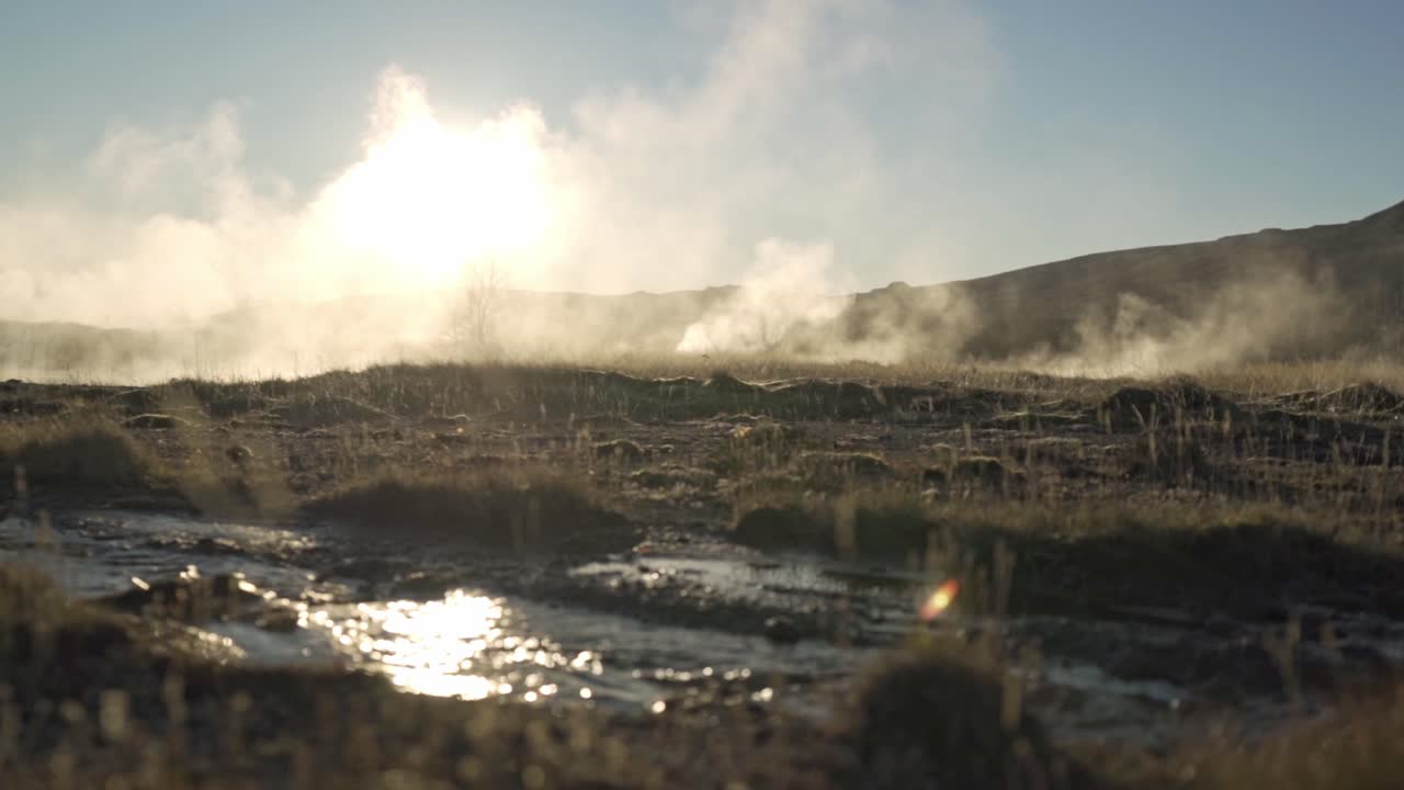 A breathtaking view of the active geothermal field at Geysir, Iceland, with powerful steam plumes backlit by the golden glow of a vibrant sunset, illuminating a rugged, barren landscape