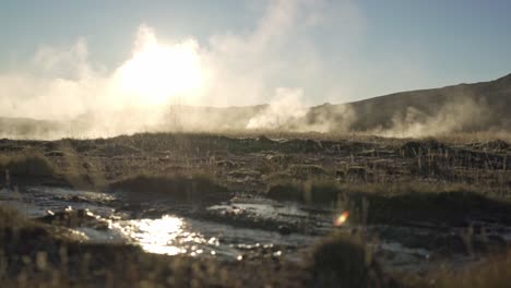 a breathtaking view of the active geothermal field at geysir, iceland, with powerful steam plumes backlit by the golden glow of a vibrant sunset, illuminating a rugged, barren landscape