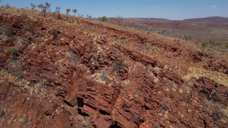 orbiting aerial shot of man climbing on red desert mountain wall during sunny day in australia - blue sky in summer with hiking adventure