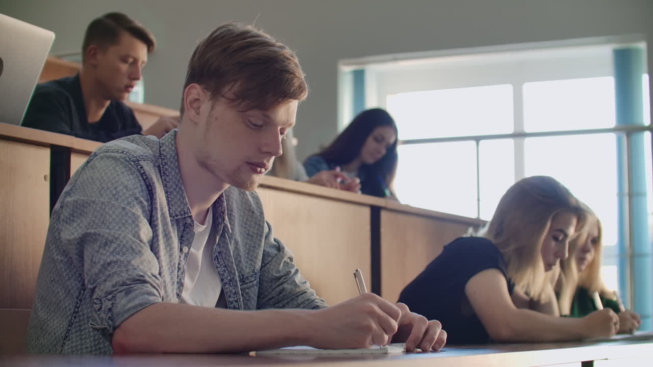 Men and women students in ordinary clothes listen to a lecture in a large audience and write a lecture notes.