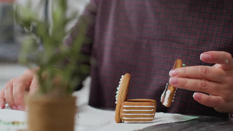 primer plano de panadero usando glaseado para decorar galletas de pan de jengibre para formar un diseño en forma de casa, con una vista borrosa de galletas de flores y otros artículos esenciales para hornear en el fondo de la cocina