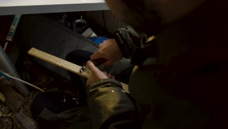 Man woodworking, carving into a long piece of wood with a small knife