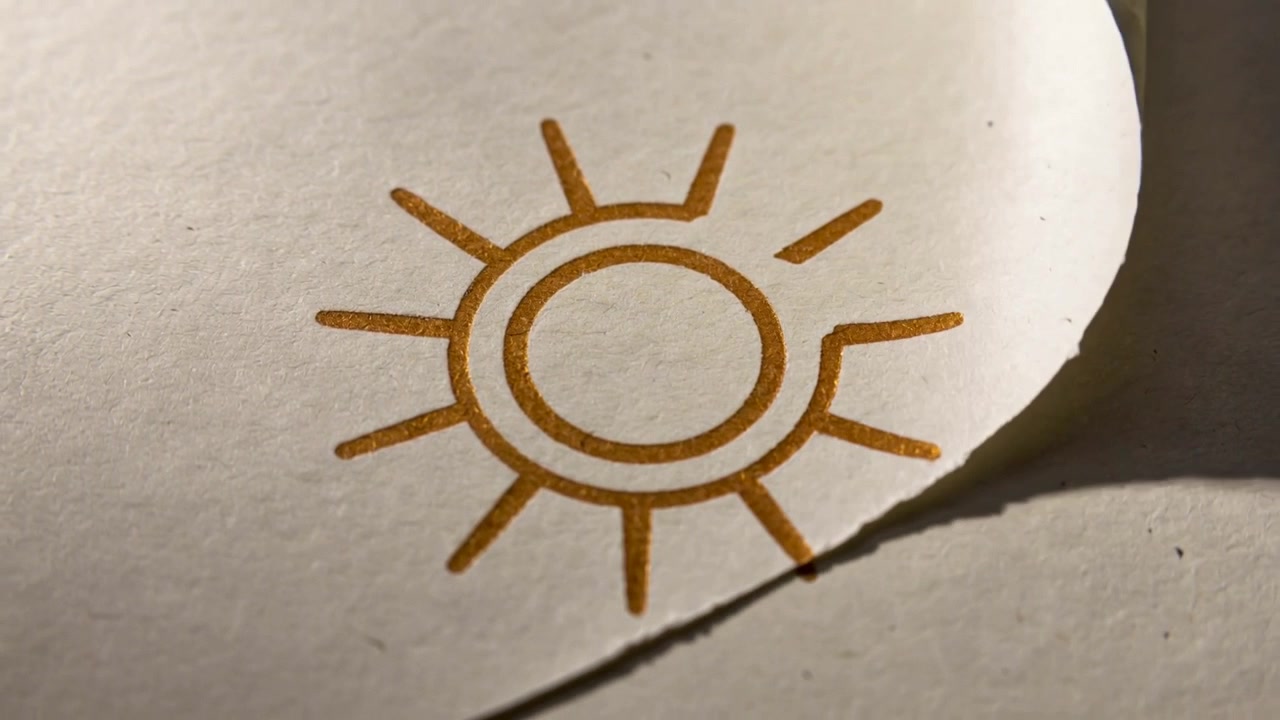 Releasing curled corner, printed sun motif unfolding on tabletop, revealing gold ink and torn edge
