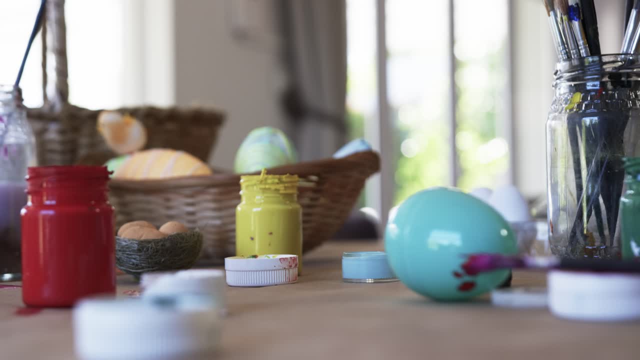 Paintbrush is leaving magenta smear on turquoise egg on wooden table near window, decorating eggs