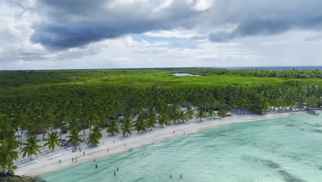 Approaching-drone-shot-flying-above-a-beachfront-located-in-the-islands-of-the-Dominican-Republic-in-the-Caribbean,-showing-a-vast-forested-area,-sandy-white-beaches-and-its-crystal-clear-waters