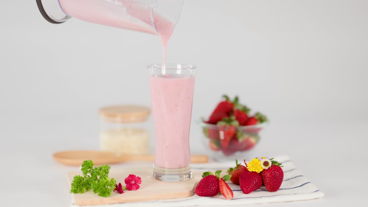 Pink strawberry milkshake poured into tall glass, surrounded by fresh strawberries, bright studio lighting