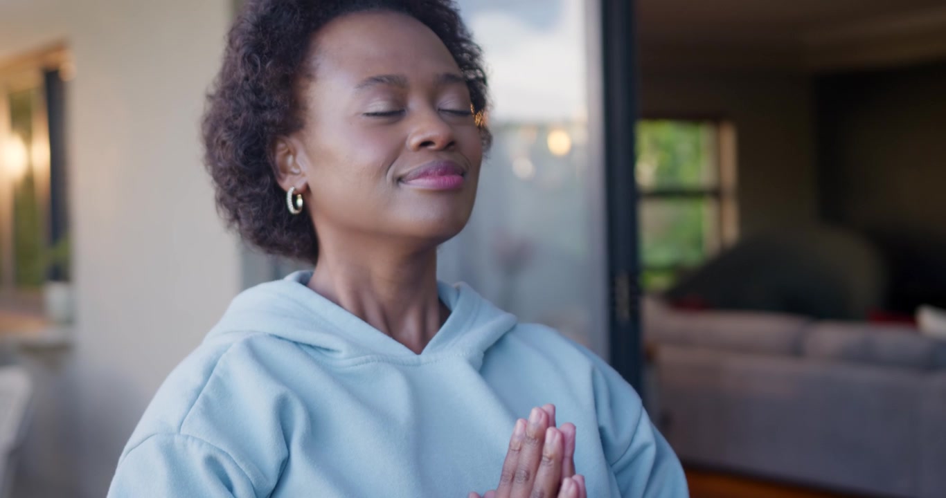 African woman pressing palms in hoodie, wristwatch at door, meditating while camera pulling back