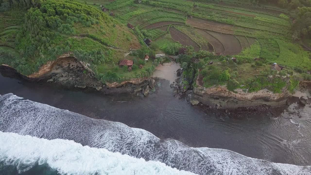 Aerial top down shot of waves reaching small private beach between cliffs and plantation fields in Indonesia