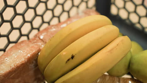 vista de cerca de una mano colocando plátanos en un carrito de compras que ya contiene un paquete de galletas, un pan y varias manzanas