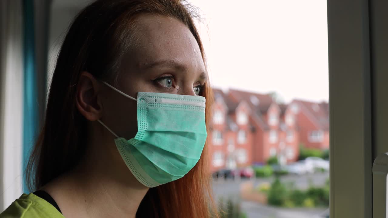 Woman with mouth nasal mask looks quarantined and looks out the window