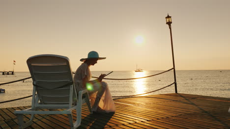 una mujer con ropa de verano se sienta en una tumbona en el muelle usa una tableta un hermoso amanecer en el mar en