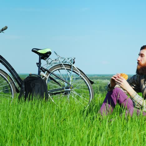 Joven-Comiendo-Una-Hamburguesa-Mientras-Está-Sentado-En-Un-Prado-Verde-Cerca-De-Su-Bicicleta