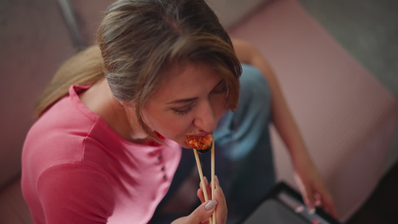 Overhead view of young adult woman in pink shirt sitting comfortably on soft couch eating sushi with chopsticks, relaxed and focused, cozy casual atmosphere with muted warm background