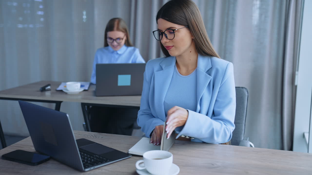 Young beautiful ladies working in the office. Long-haired ladies wearing glasses sit at the desk at their laptops. Blurred backdrop.