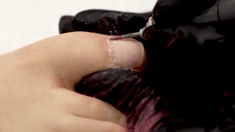 female manicurist removes cuticle on big toe with nail drill, performs pedicure for woman customer using different nozzle of electric pedicure machine