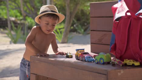 young latin baby boy playing with his car toys on a bench covered in sand at the beach on a sunny summer day wearing a hat