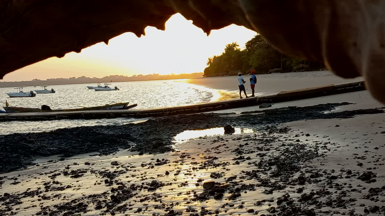 puesta de sol en la playa de la isla vista desde debajo de un tronco de árbol y con pequeños botes en el fondo
