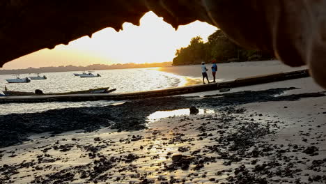 Sunset-on-the-beach-of-the-island-seen-from-under-a-tree-trunk-and-with-small-boats-in-the-background