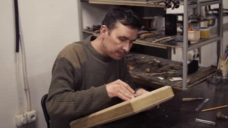 caucasian joiner makes notches on the wooden workpiece with curved chisel. a craftsman carving wooden ornament on backgammon sitting in his working place. slow motion