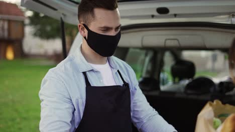 Female-customer-is-getting-paper-bag-of-organic-groceries-from-male-courier-in-a-mask,-apron-and-gloves