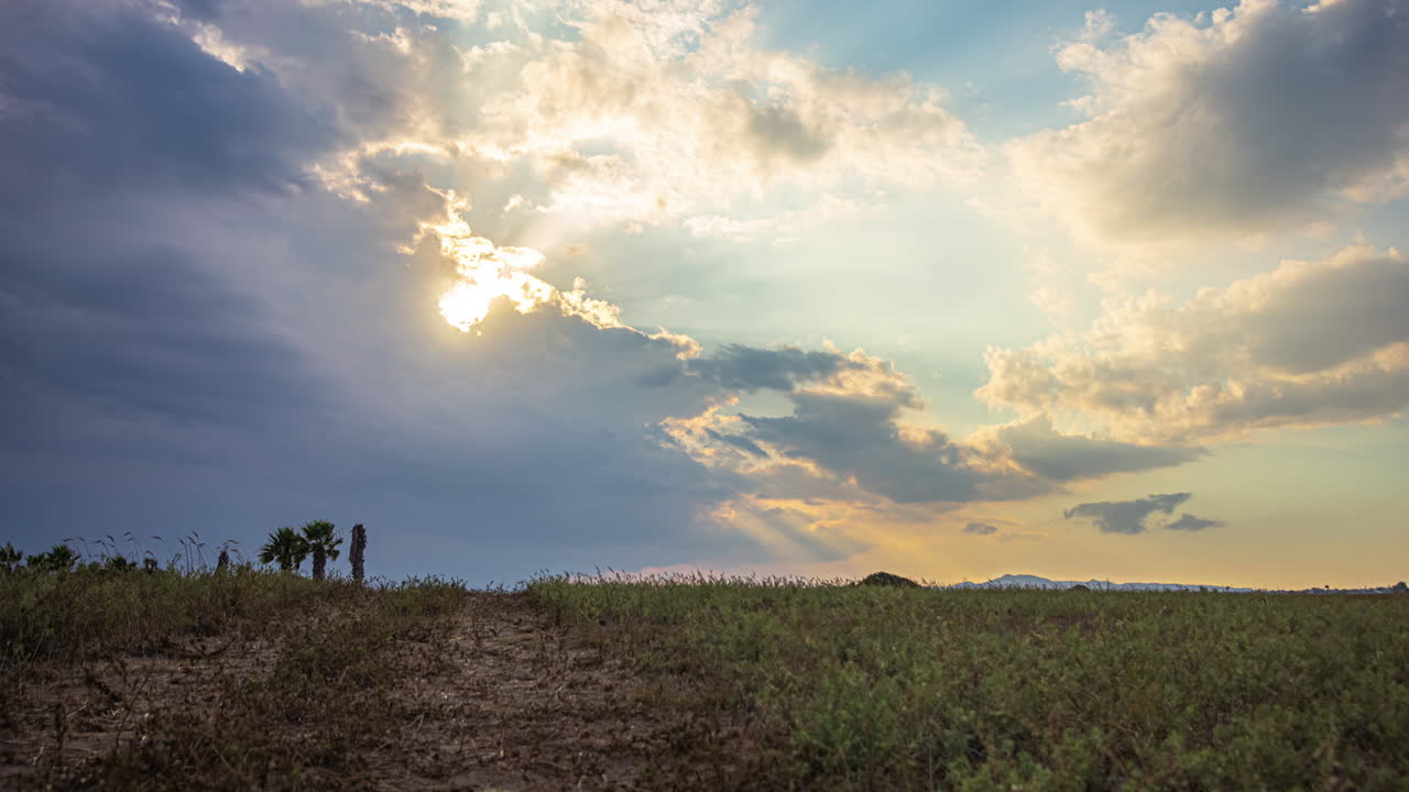 Filmato timelapse cinematografico di nuvole in movimento e il sole che fa capolino tra le nuvole, con un campo d'erba e alcuni alberi in primo piano.