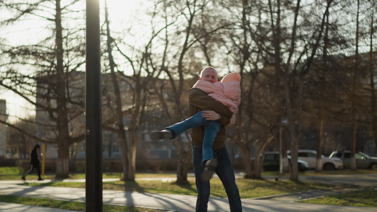 мужчина в коричневом пиджаке и синих джинсах с радостью носит и вращает маленькую девочку в розовой шапке и пиджаке в освещенном солнцем парке.