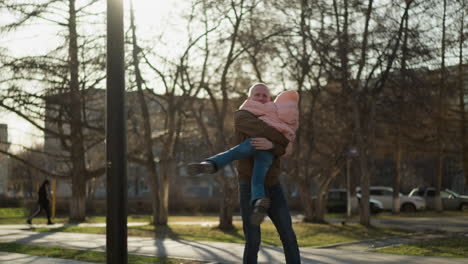 a man wearing a brown jacket and blue jeans joyfully carrying and spinning a little girl in a pink cap and jacket in a sunlit park. a playful and heartwarming moment of father-daughter bonding